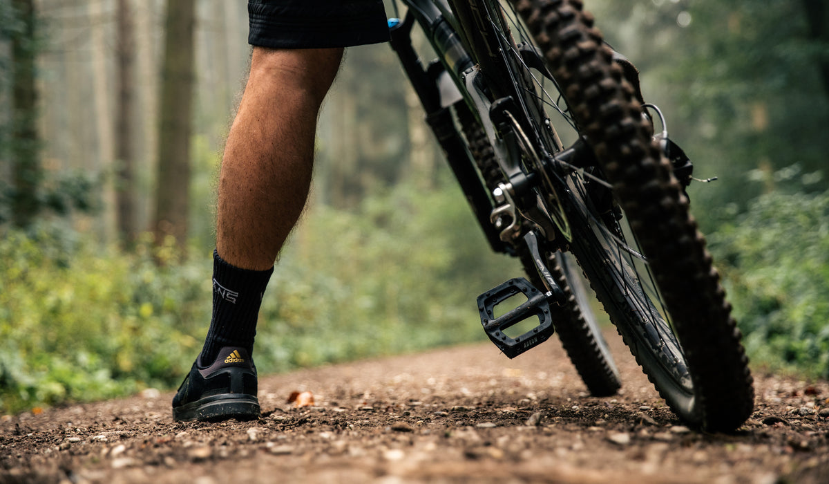 Ein Radfahrer steht mit einem Fuß auf dem Boden und einem auf dem Pedal und hält ein Mountainbike auf einem unbefestigten Weg in einem Wald. Die untere Hälfte des Radfahrers und der Reifen des Fahrrads sind sichtbar.