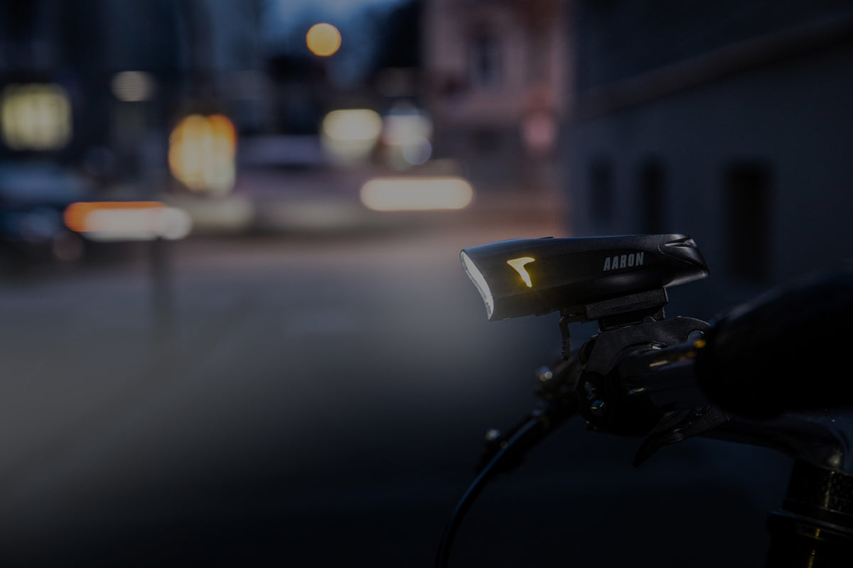Ein Fahrradlenker mit einem hellen Scheinwerfer beleuchtet in der Dämmerung eine schummrige, verschwommene Straßenszene. Der Name AARON ist auf der Lampe zu lesen.