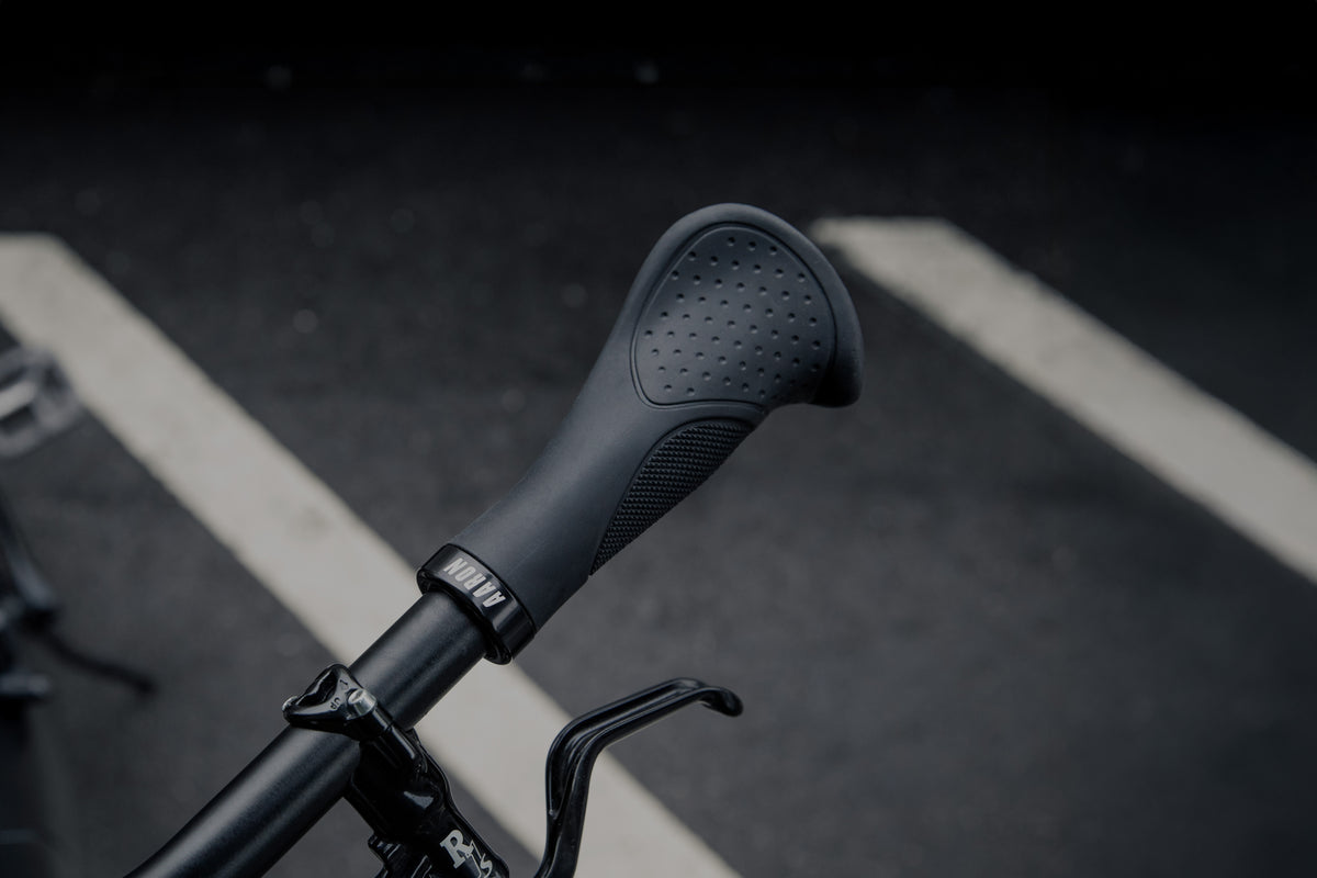 Nahaufnahme eines schwarzen ergonomischen Fahrradlenkergriffs mit strukturierter Oberfläche, der über einer gepflasterten Straße angebracht ist, wobei im Hintergrund zwei weiß gestrichene Linien zu sehen sind.