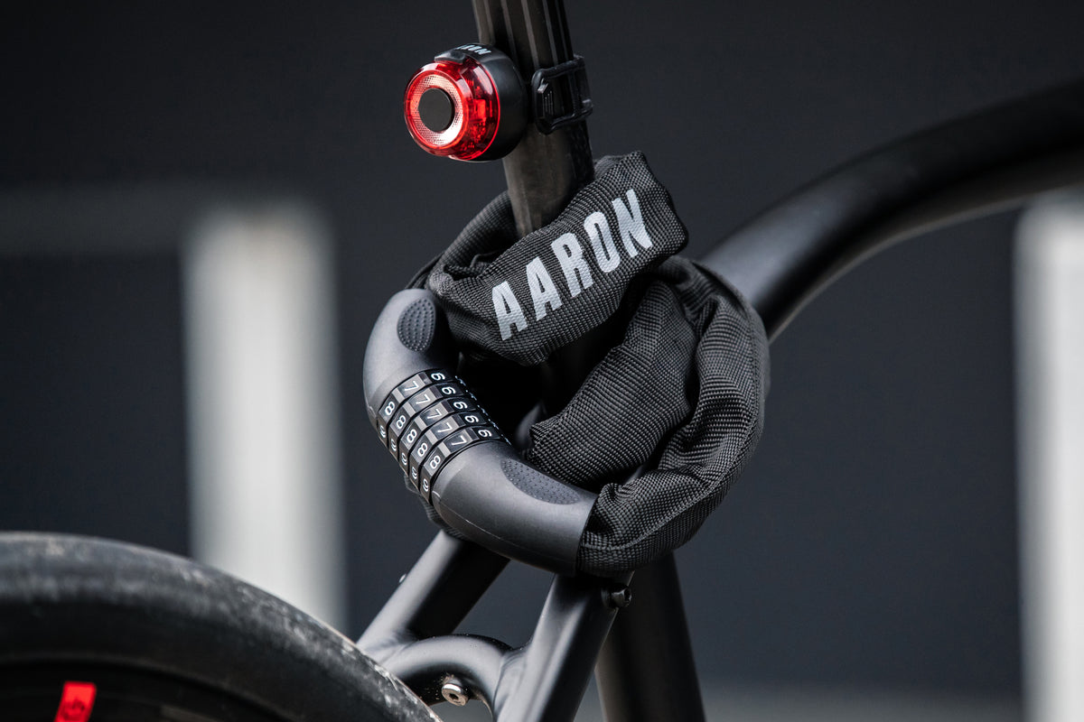 Ein Fahrrad ist mit einem schwarzen Zahlenschloss gesichert, das in eine Schutzhülle mit der Aufschrift AARON eingepackt ist. Ein rotes Rücklicht ist an der Sattelstütze befestigt. Der Hintergrund ist unscharf und dunkel.