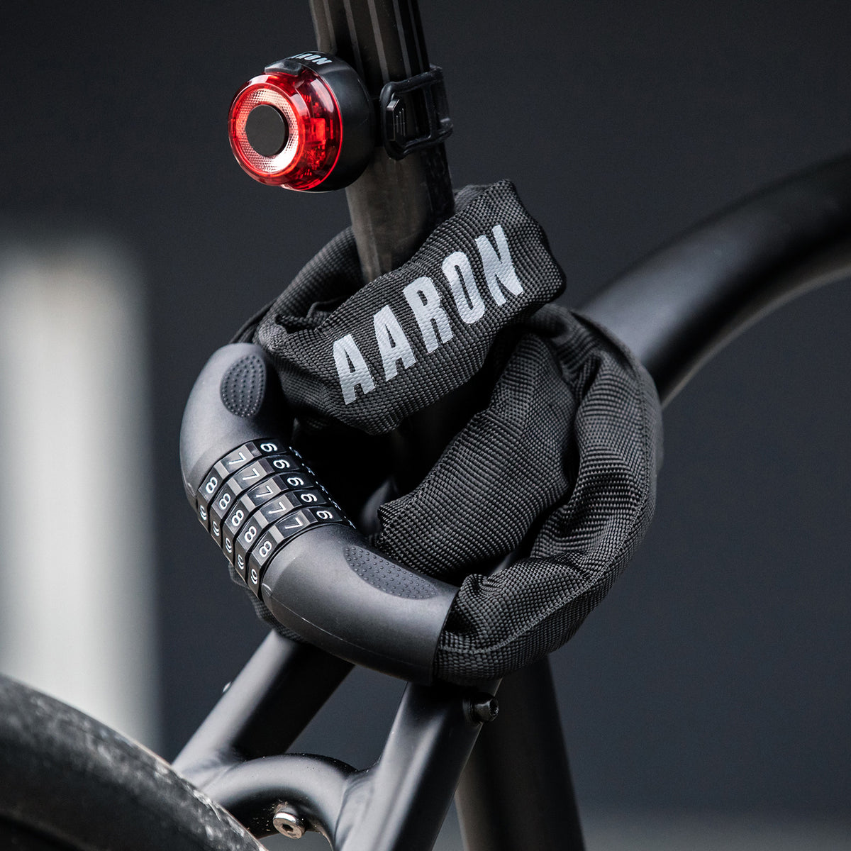Ein schwarzes Fahrradschloss mit einer Stoffabdeckung mit der Aufschrift AARON sichert einen Fahrradrahmen. Ein rotes Fahrradrücklicht ist an der Sattelstütze befestigt, und der Hintergrund ist unscharf.