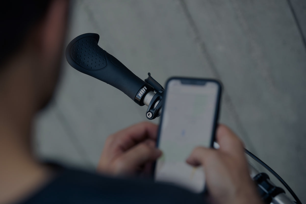 Eine Person, die ein Smartphone in einer Hand hält und mit der anderen einen Fahrradlenker umgreift, von der Schulter aus gesehen, wobei der Bildschirm des Telefons leicht verschwommen ist.