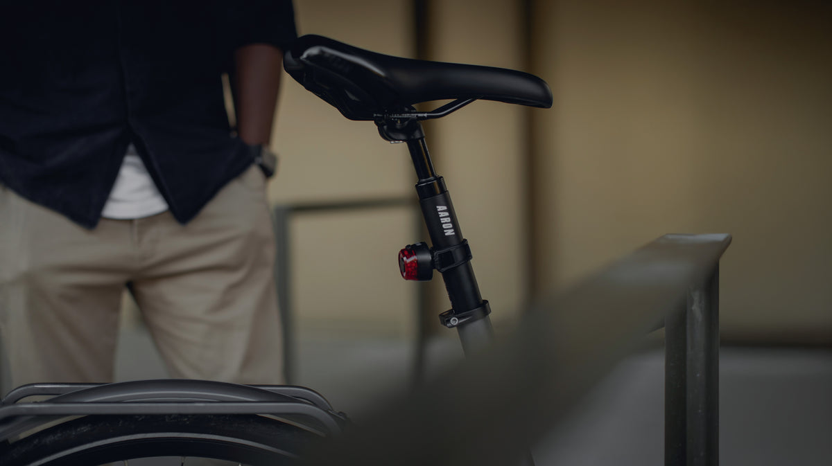 Eine Person steht hinter einem Fahrradsattel, teilweise sichtbar mit den Händen in den Taschen, und trägt ein dunkles Hemd und eine beigefarbene Hose. Der Fokus liegt auf der Fahrradsattelstütze und dem angebrachten Rücklicht.