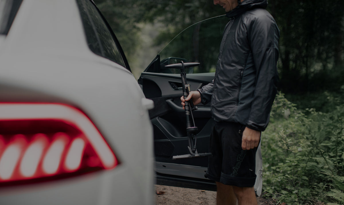 Eine Person in Outdoor-Kleidung steht neben einer offenen Autotür und hält eine tragbare Luftpumpe in der Nähe eines weißen Fahrzeugs in einem Waldgebiet.
