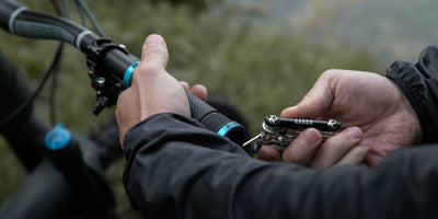 Nahaufnahme einer Person in dunkler Jacke, die einen Fahrradlenker mit einem Multitool einstellt, mit unscharfem Grün im Hintergrund.