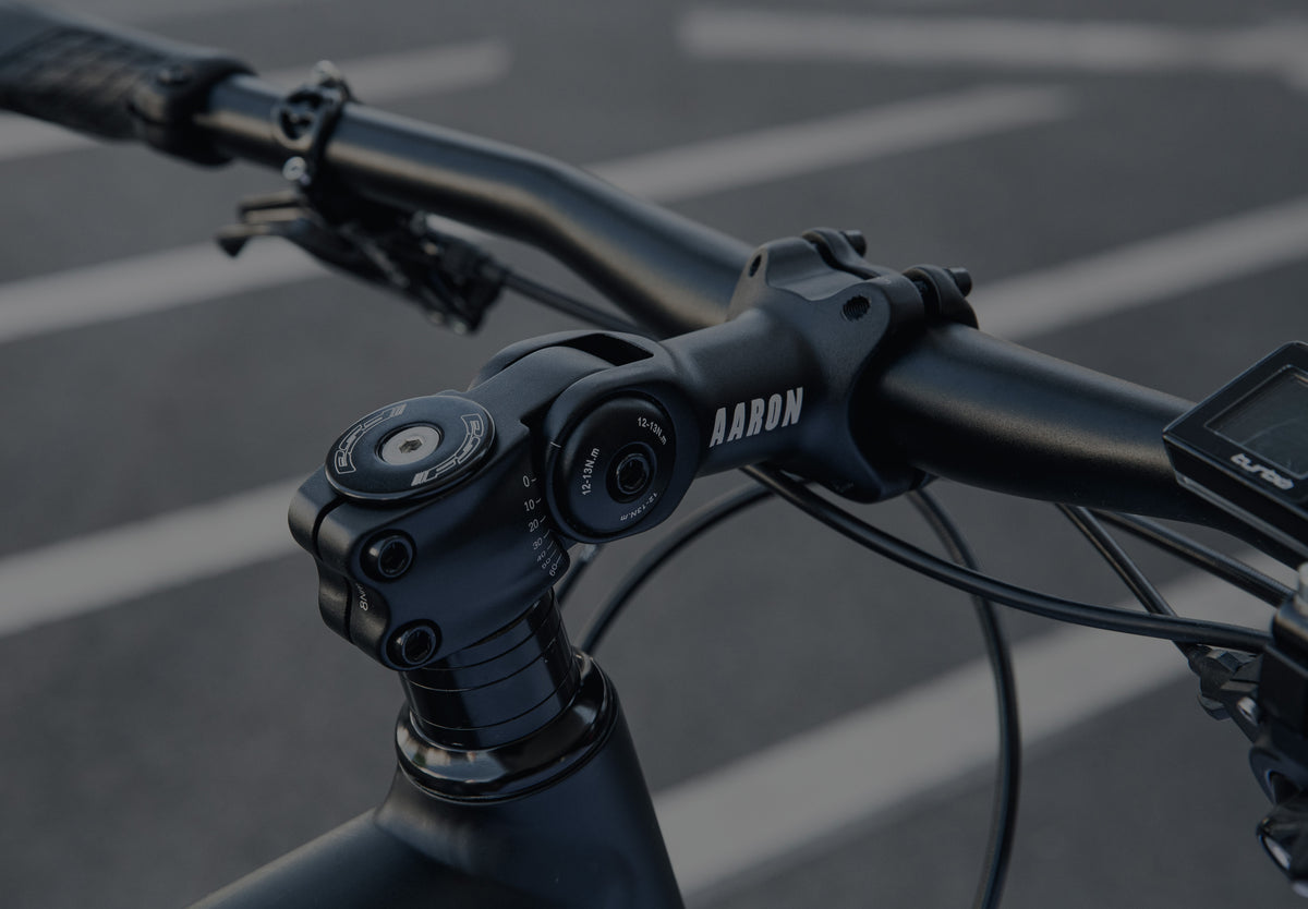 Nahaufnahme eines schwarzen Fahrradlenkers und Vorbaus mit Einstellrädern und dem Markennamen "AARON". Der Hintergrund zeigt einen verschwommenen Bürgersteig mit weiß aufgemalten Linien.