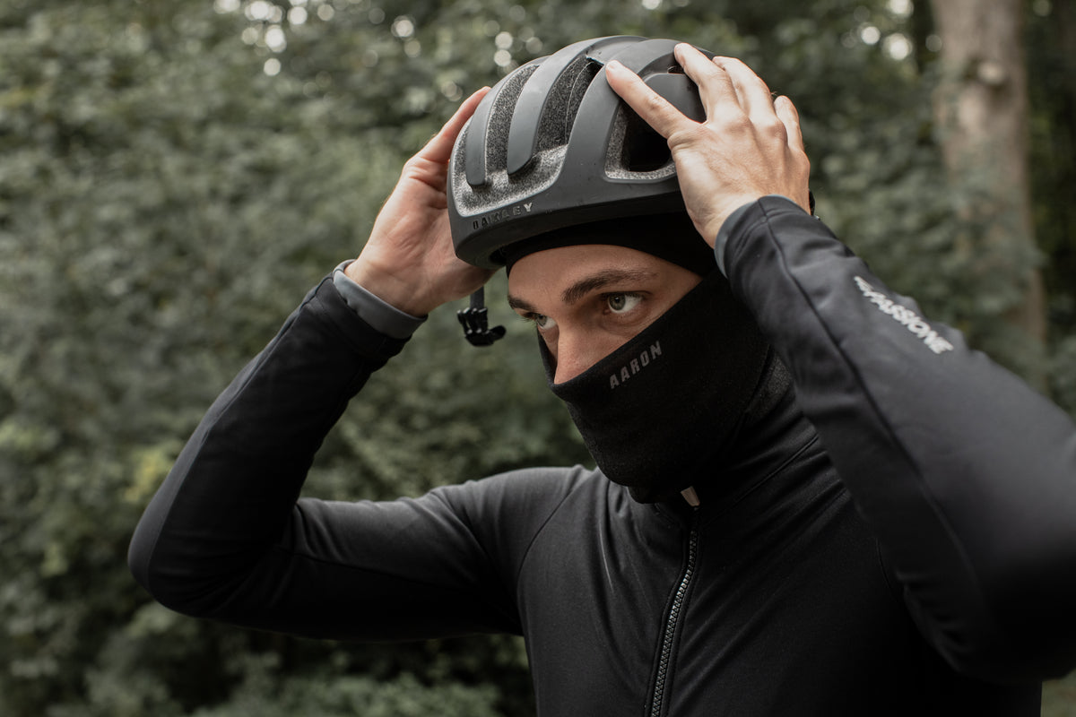Eine Person, die eine schwarze Sturmhaube und eine schwarze Jacke trägt, passt einen schwarzen Fahrradhelm im Freien an, mit einem Hintergrund aus grünen Bäumen.