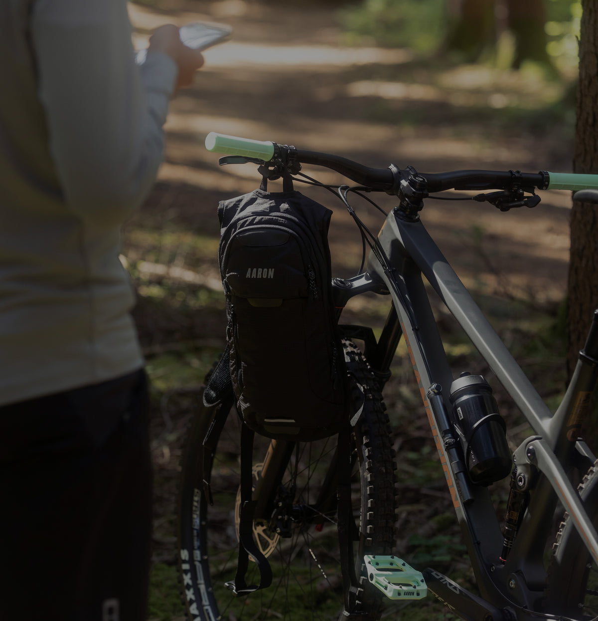 Eine Person steht in einem Wald mit einem Smartphone in der Hand neben einem Mountainbike, an dessen Lenker ein schwarzer Rucksack mit der Aufschrift AARON befestigt ist. Das Sonnenlicht fällt durch die Bäume auf den unbefestigten Weg.