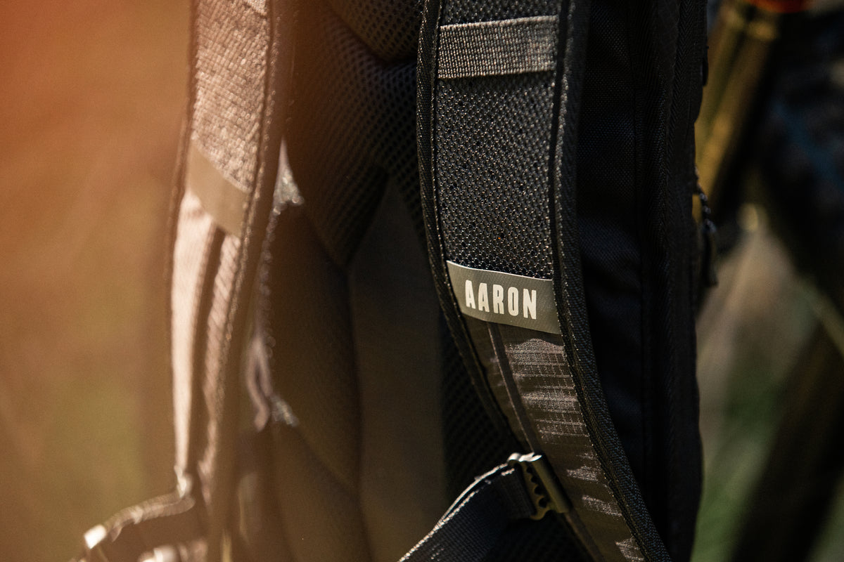 Nahaufnahme eines schwarzen Rucksackträgers mit einem Namensschild mit der Aufschrift AARON, vor einem unscharfen Hintergrund im Freien mit warmem Sonnenlicht.
