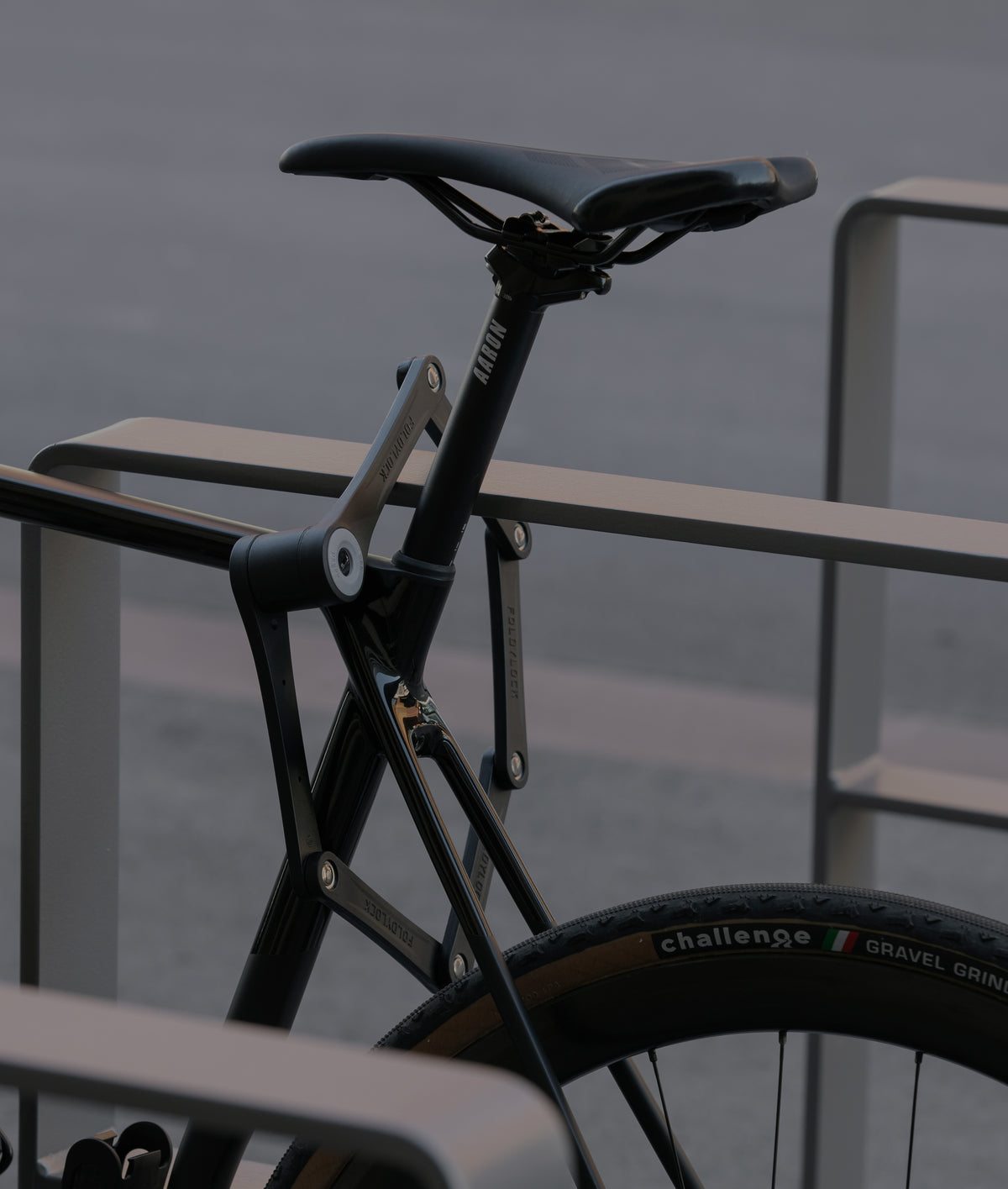 Nahaufnahme eines schwarzen Fahrrads, das an einem Metallgepäckträger befestigt ist, wobei der Schwerpunkt auf dem Sattel, dem Rahmen und dem Hinterrad liegt, auf dem ein GRAVEL GRINDER Reifen zu sehen ist. Der Hintergrund ist ein verschwommener Bürgersteig.