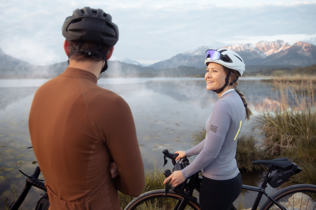 Zwei Radfahrer mit Helmen und Sportkleidung stehen neben ihren Fahrrädern an einem ruhigen See, mit Bergen und Nebel im Hintergrund. Die Frau lächelt und schaut den Mann an.