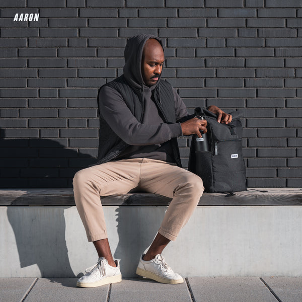 AARON Rolltop Rucksack für Damen und Herren - Schwarz mit Mann auf Bank beim Öffnen des Rucksacks