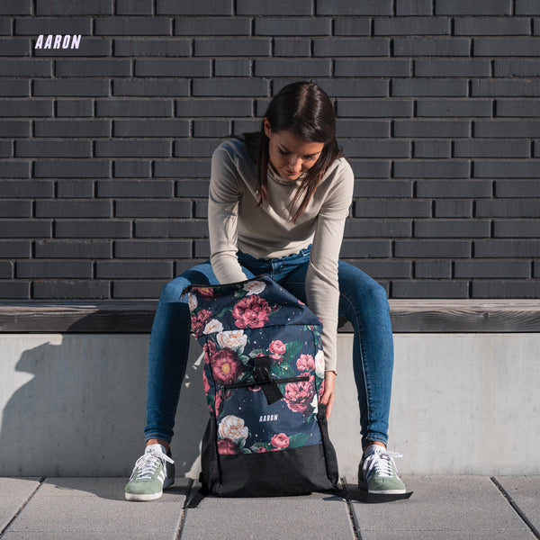 Frau sitzt draußen und hält den AARON Rolltop 2.0 Rucksack für Damen und Herren mit Blumenmuster auf dem Boden.