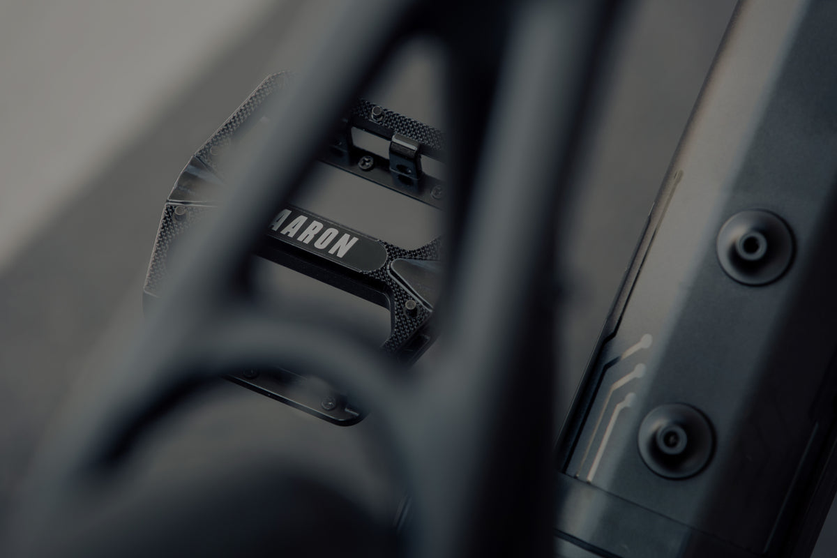 Nahaufnahme eines schwarzen Fahrradpedals mit dem aufgedruckten Namen AARON, teilweise verdeckt durch den Rahmen des Fahrrads und fotografiert bei gedämpfter, gedämpfter Beleuchtung.