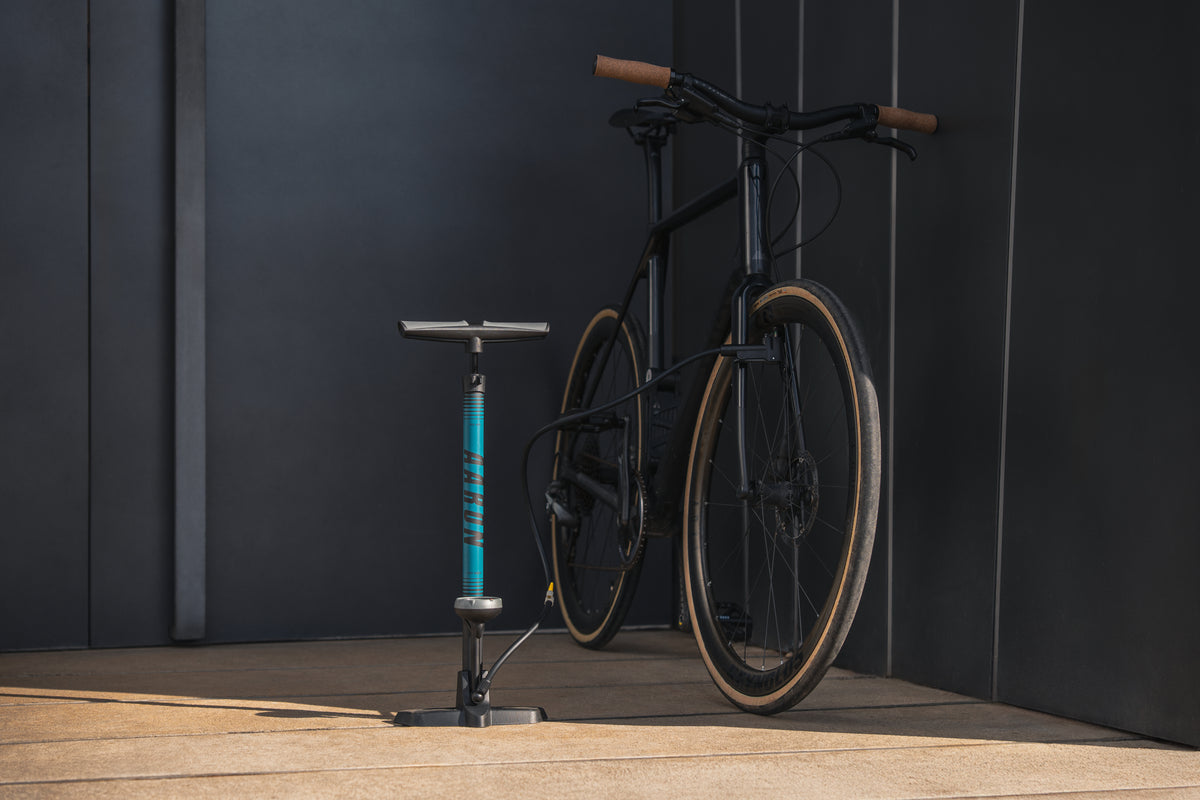 Ein schwarzes Fahrrad mit hellbraunen Reifen ist vor einer dunklen Wand auf einer Betonfläche geparkt. Eine blau-schwarze Standradpumpe steht aufrecht vor dem Fahrrad.