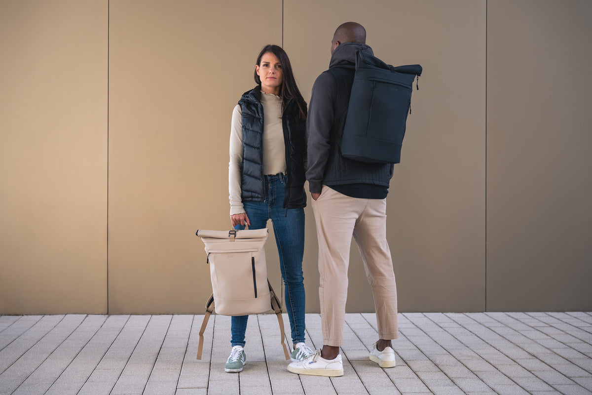 Zwei Personen stehen sich vor einer einfarbig beigen Wand gegenüber. Die eine hat einen beigen Rucksack in der Hand, die andere trägt einen schwarzen Rucksack. Beide sind leger mit Jacken, Jeans und Turnschuhen bekleidet.
