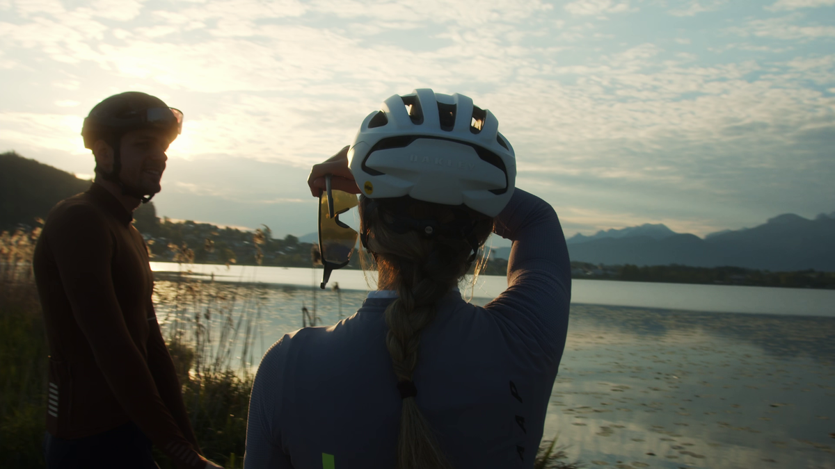 Zwei Radfahrer mit Helmen stehen bei Sonnenuntergang an einem Seeufer. Eine Person, die von hinten mit einem Zopf zu sehen ist, hebt das Fernglas, um über das Wasser zu blicken, während die andere ihnen gegenübersteht. Im Hintergrund sind Berge zu sehen.